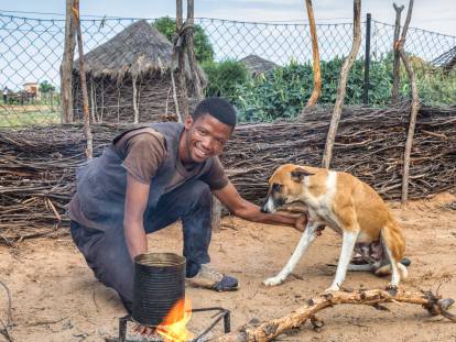 Un homme africain en train de cuisiner au dehors, avec un chien à ses côtés