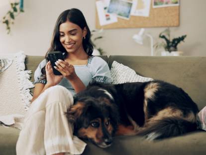 Une femme assise sur un canapé qui regarde son téléphone avec un chien couché à côté d'elle sur le canapé