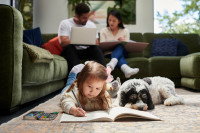 Une petite fille allongée par terre aux côtés de son chien, avec ses parents assis sur un canapé en arrière-plan