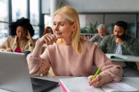 Une femme en salle de classe qui regarde son ordinateur tout en écrivant sur son cahier