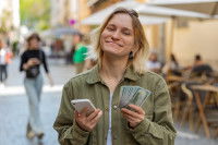 Une femme debout dans la rue tenant son téléphone dans une main et des dollars dans l'autre