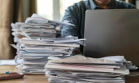 Une personne en train d'utiliser son ordinateur portable avec des piles de papiers à côté d'elle
