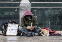 Un homme sans-abri assis par terre, entouré de ses affaires et de son chien, avec un panneau sur lequel il demande de l'argent