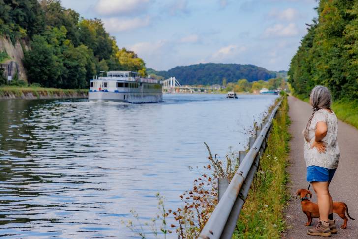 Un Teckel tenu en laisse par une femme, au bord d’une rivière en Belgique, observant un bateau au loin.