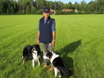 Deux Border Collies au pied d'un homme portant le gilet et la casquette de l'AFBC dans une prairie verdoyante