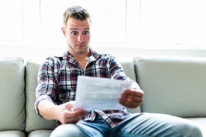 Un homme assis sur le canapé lisant une feuille de papier, avec une expression choquée