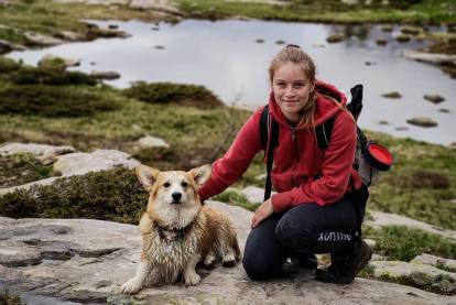 Un Welsh Corgi Pembroke allongé sur une montagne à côté d'une femme accroupie et portant un sac au dos